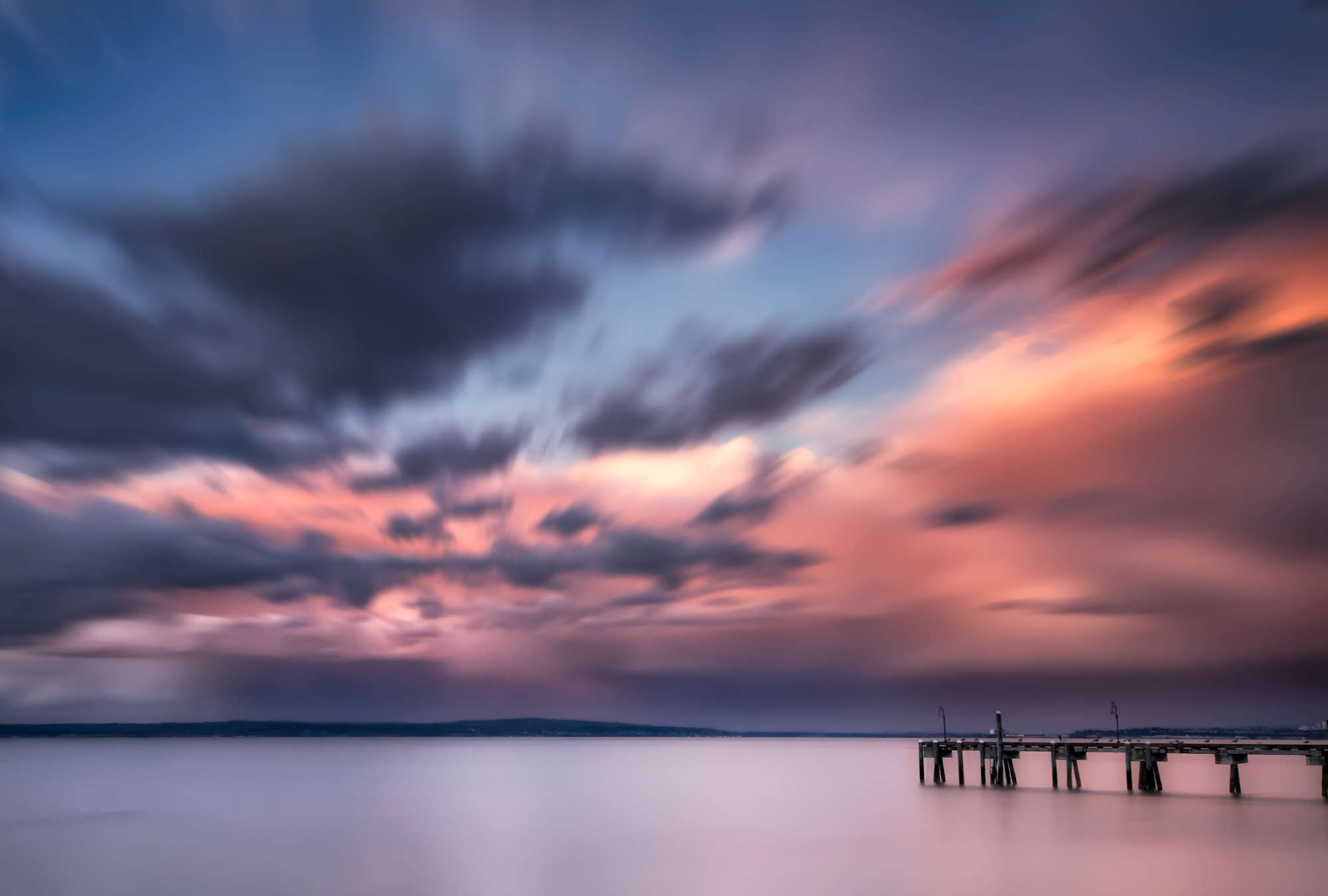             Fototapete Küstenlandschaft mit Steg im Wasser und buntem Wolkenspiel
        